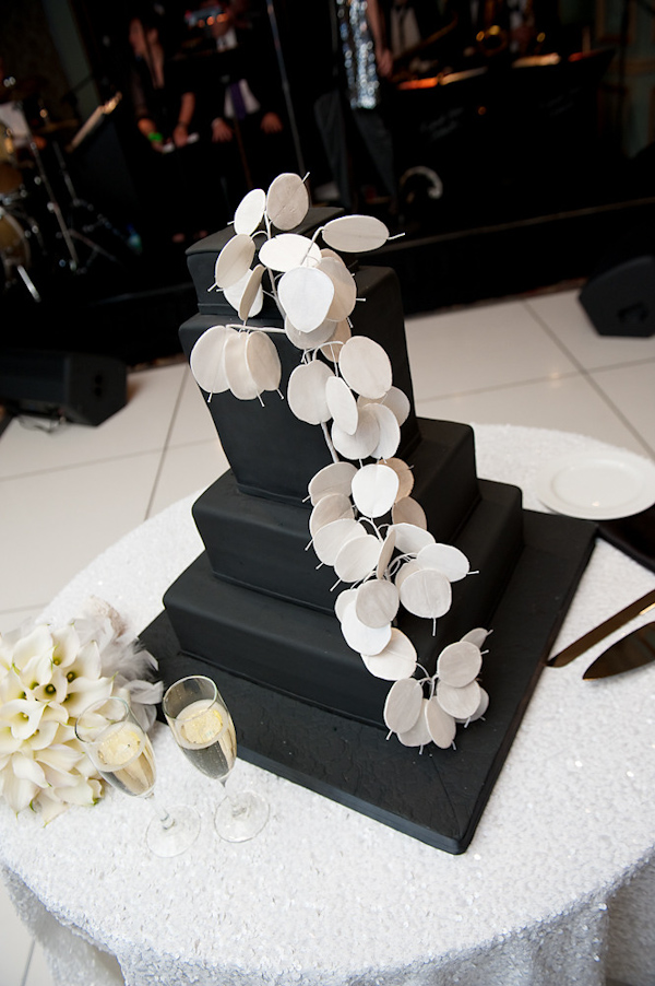 black four tiered square wedding cake on a black cake stand with white design draping down cake as topper - cake is on round table with white table cloth with bride and groom's champagne flutes and an ivory floral arrrangement -  photo by Houston based wedding photographer Adam Nyholt